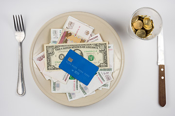 Image showing On the plate is a lot of money and a plastic credit card, standing next to a glass and a fork and knife lie, top view