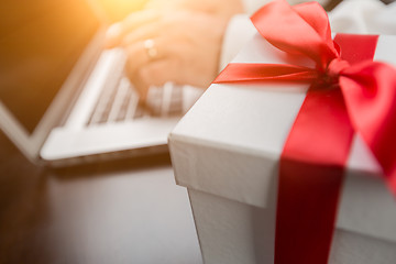 Image showing White Gift Box with Red Ribbon and Bow Near Man Typing on Laptop