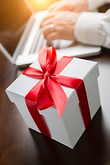 Image showing White Gift Box with Red Ribbon and Bow Near Man Typing on Laptop