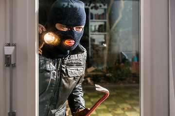 Image showing Masked burglar with flashlight and crowbar looking into glass wi