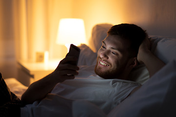 Image showing happy young man with smartphone in bed at night