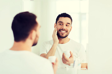 Image showing happy young man applying cream to face at bathroom