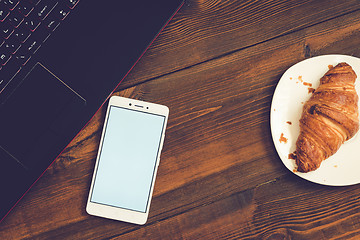 Image showing Workspace with laptop, smartphone and croissant on wooden desk