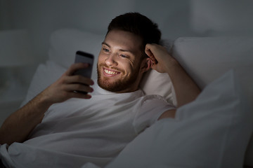 Image showing happy young man with smartphone in bed at night