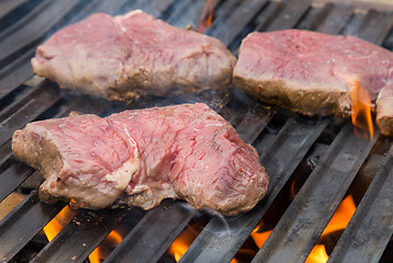 Image showing Raw beef steaks on grill with flames.