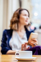 Image showing cup of tea on the table and a woman with a smartphone in the blu