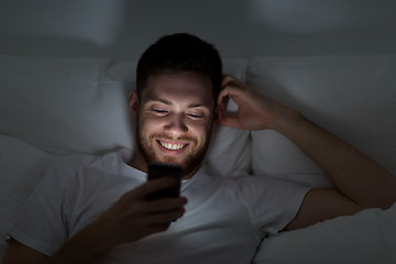 Image showing happy young man with smartphone in bed at night