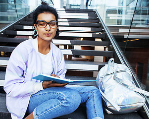Image showing young cute indian girl at university building sitting on stairs reading a book, wearing hipster glasses, lifestyle people concept