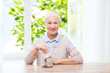 Image showing senior woman putting money into glass jar at home