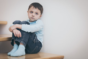 Image showing one sad little boy sitting on the stairs in house at the day tim