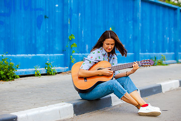 Image showing woman in jeans sits on a road curb and plays the guitar