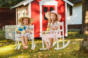 Image showing Cute little blond girls in hats sitting on the field with soft toys in summer.