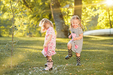 Image showing The cute little blond girls in rubber boots playing with water splashes on the field in summer