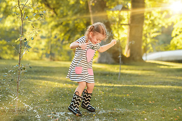 Image showing The cute little blond girl in rubber boots playing with water splashes on the field in summer