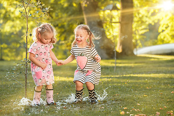 Image showing The cute little blond girls in rubber boots playing with water splashes on the field in summer