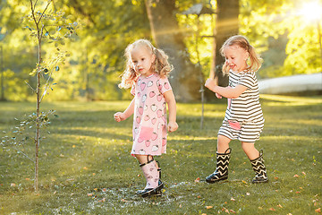 Image showing The cute little blond girls in rubber boots playing with water splashes on the field in summer