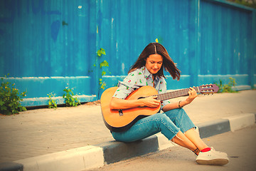 Image showing woman in jeans sits on a road curb and plays the guitar