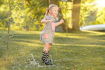 Image showing The cute little blond girl in rubber boots playing with water splashes on the field in summer