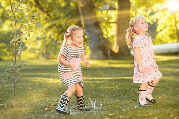 Image showing The cute little blond girls in rubber boots playing with water splashes on the field in summer