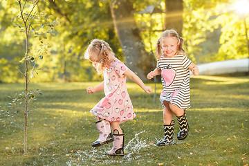 Image showing The cute little blond girls in rubber boots playing with water splashes on the field in summer