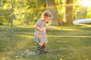 Image showing The cute little blond girl in rubber boots playing with water splashes on the field in summer