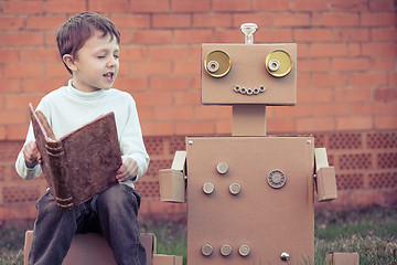 Image showing One little boy reading to  robot from cardboard boxes outdoors.