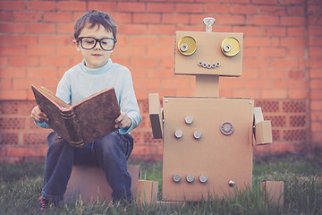 Image showing One little boy reading to  robot from cardboard boxes outdoors.