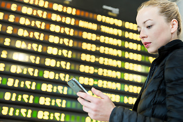 Image showing Woman at airport in front of flight information board checking her phone.