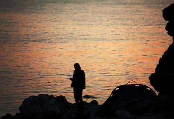 Image showing Silhouette of trekker on rocky seashore near camping tent on ove