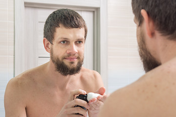 Image showing An unshaven man applies a shaving foam on the palm of his hand, and looks in the mirror in the bathroom