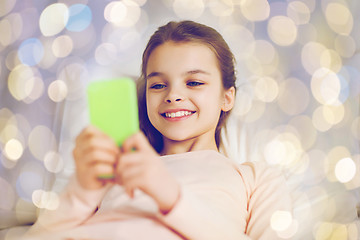 Image showing happy girl in bed with smartphone over lights