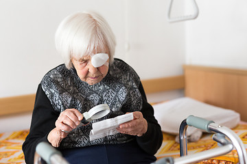 Image showing Visually impaired elderly 95 years old woman sitting at the bad trying to read with magnifying glass.