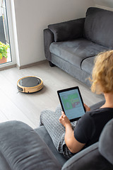 Image showing Woman is steering her vacuum cleaning robot from the sofa