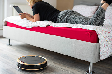 Image showing Vacuum cleaning robot with relaxed woman reading in the background