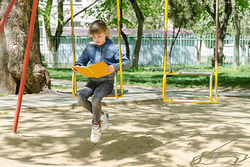 Image showing A girl on a walk reading a book swinging on a swing at the playground
