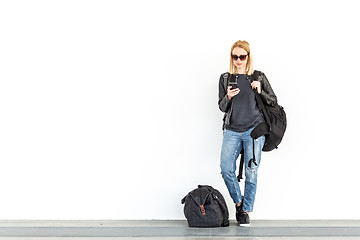 Image showing Fashionable young woman using her mobile phone while standing and waiting against plain white wall on the station whit travel bag by her side.