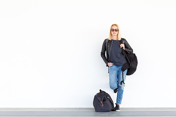 Image showing Fashionable young woman standing and waiting against plain white wall on the station whit travel bag by her side.