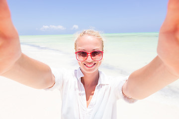 Image showing Young woman wearing white beach tunic taking selfie on tropical white sandy beach.