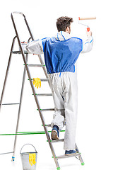 Image showing Young male decorator painting with a paint roller climbed a ladder isolated on white background.