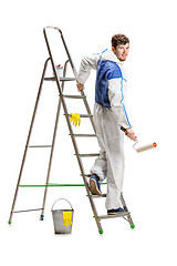 Image showing Young male decorator painting with a paint roller climbed a ladder isolated on white background.