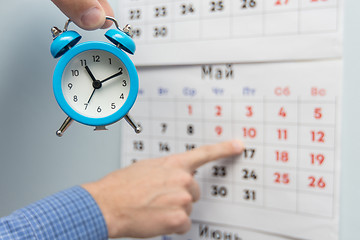 Image showing A hand holds a small alarm clock, in the background a hand points to long weekends and holidays on a wall calendar