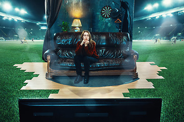 Image showing Boring fan is sitting on the sofa and watching TV in the middle of a football field.