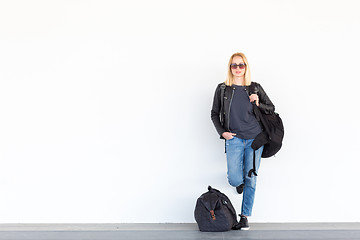 Image showing Fashionable young woman standing and waiting against plain white wall on the station whit travel bag by her side.