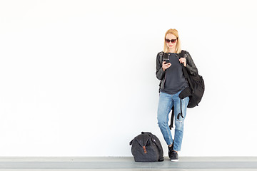 Image showing Fashionable young woman using her mobile phone while standing and waiting against plain white wall on the station whit travel bag by her side.