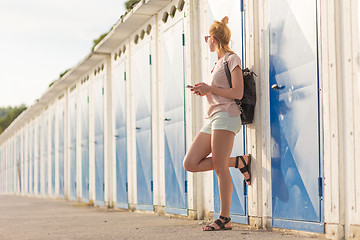 Image showing Blonde young female traveler wearing summer style clothing, holding mobile phone, against retro blue beach dressing rooms at summer time vacation.