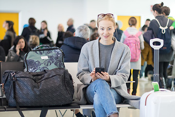 Image showing Cheerful female traveler smiling, looking at camera while reading on her cell phone while waiting to board a plane at departure gates at airport terminal.