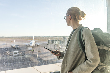 Image showing Casually dressed female traveler at airport, holding smart phone device, looking through the airport gate windows at planes on airport runway