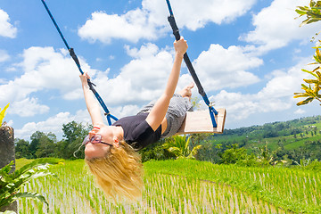 Image showing Happy female traveller swinging on wooden swing, enjoying summer vacation among pristine green rice terraces.