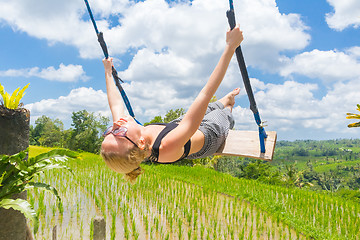 Image showing Happy female traveller swinging on wooden swing, enjoying summer vacation among pristine green rice terraces.