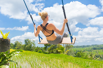 Image showing Happy female traveller swinging on wooden swing, enjoying summer vacation among pristine green rice terraces.